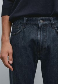 Mörkblå denimjeans med knäppt midja, fem fickor och kontrasterande sömmar. Tyget har en slät, något texturerad yta.