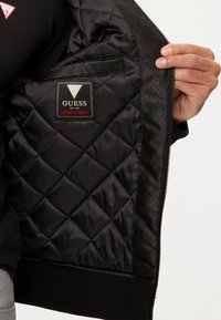 Zwarte gewatteerde jas interior met een "GUESS" label. Soepele stof met een ruitpatroon; ribgebreide manchetten en zoom, met ritsluiting.