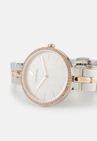 Reloj de plata y oro rosa con un dial texturizado blanco, bisel acentuado con cristales y una pulsera metálica de eslabones con acentos dorados.
