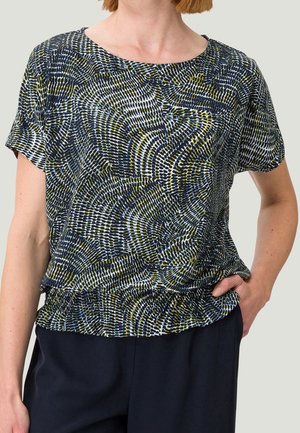 Femme portant un chemisier à manches courtes avec un motif circulaire abstrait noir, blanc et olive, rentré dans un pantalon foncé, main posée sur la hanche.