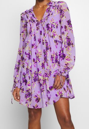 Damesjurk met lange mouwen in lavendel, voorzien van een bloemenprint in paars en geel, plooidetails en aantrekkoorden bij de manchetten, net boven de knie vallend.