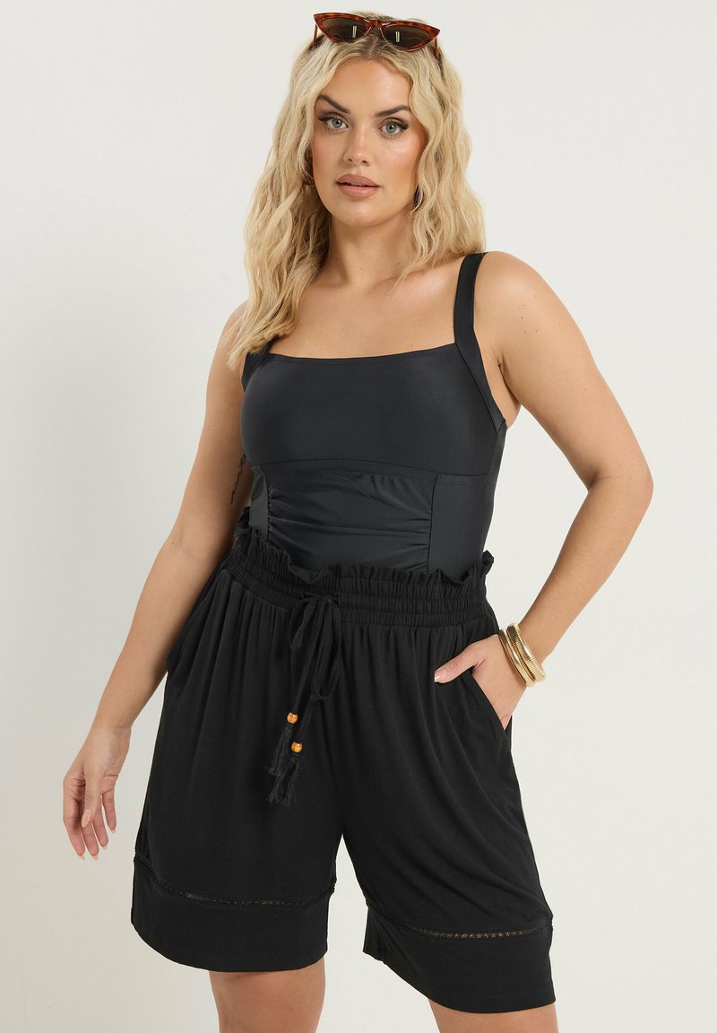 Blonde vrouw met zonnebril op haar hoofd, gekleed in een zwart mouwloos topje en zwart, hooggetailleerd short met trekkoord, staand voor een beige achtergrond.