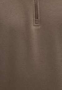 Brown zip-up garment with a high collar, featuring a textured surface and a contrasting zipper. Material appears soft and stretchy.