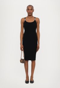 Rochie neagră cu dungi, cu bretele ajustabile accentuate cu argintiu, siluetă mulată și design până la genunchi. Modelul ține o geantă mică.