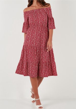 Kvinde iført en rød off-shoulder midi-kjole med hvidt blomsterprint og klokkeærmer, kombineret med hvide hælede sandaler med stropper.