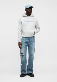 Mladić u svijetlosivoj hoodici s natpisom "Sounds gay I'm in", poderanim plavim trapericama, crnim cipelama i svijetloplavom kapom, stoji na bijeloj pozadini.