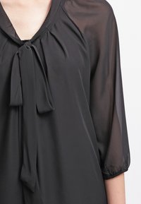 Zwarte doorschijnende blouse met een V-hals, gestrikte strikdetail bij de kraag en driekwartmouwen verzameld bij de manchetten. Zacht, lichtgewicht stof.