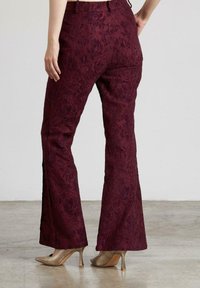 Pantalones acampanados de tejido burdeos profundo con un patrón floral texturizado. Presentan una línea de cintura y aberturas en los tobillos. Combinados con zapatos beige.