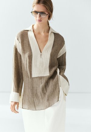 Kvinde iført brun og hvid langærmet bluse med bred krave og hvide bukser, accessoriseret med store aviator-solbriller.