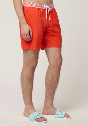 Shorts de bain orange vif avec une taille élastique et des rayures rouges et blanches. Un cordon de serrage et des poches latérales ajoutent de la fonctionnalité.