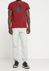 Punainen lyhythihainen t-paita, jossa on musta Adidas-logo, yhdistettynä vaaleanharmaisiin kapeisiin jogger-housuihin ja mustiin urheilukenkiin, joissa on valkoiset pohjat.