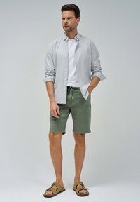 Camisa ligera de rayas, tipo botón, sobre una camiseta blanca, combinada con pantalones cortos de color verde oliva y sandalias marrones. Ajuste simple y casual.