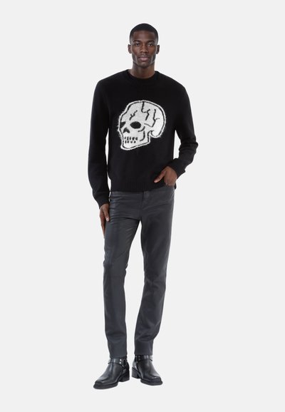 Suéter de punto negro con un gráfico de calavera blanca en el frente, combinado con pantalones negros ajustados y botas de tobillo con detalle de hebilla.