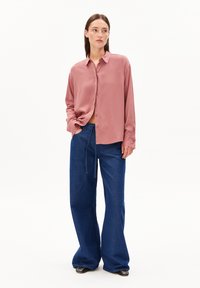 Woman wearing loose pink button-up shirt, wide-leg blue jeans with tie belt, and black studded pointed shoes, standing with one hand in pocket.