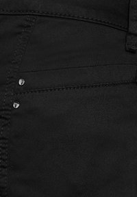 Tessuto denim nero con una superficie liscia, caratterizzato da due borchie metalliche sopra una tasca frontale e cuciture dettagliate lungo le cuciture.