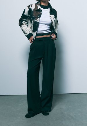 Kurtka ze sztucznego futra w brązowo-biały wzór w cętki krowy, noszona na czarnej bluzce, zestawiona z ciemnymi szerokimi spodniami i czarnymi butami.