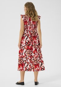 Robe maxi avec un buste froncé, des volants aux épaules et un motif floral rouge, rose et blanc éclatant sur fond noir. Des chaussures plates noires sont portées.