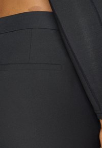 Imagem de perto de uma pessoa a usar calças pretas sob medida com um bolso traseiro com costura e uma blusa preta de mangas longas visível.