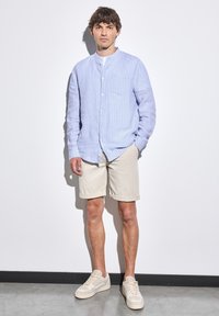 Lichtblauw gestreept overhemd met een mandarinakraag, gecombineerd met beige shorts en schoenen in neutrale tinten op een grijze vloer.