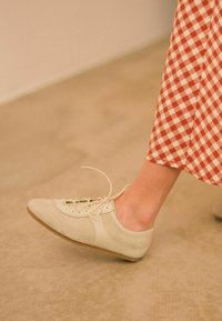 Scarpa in suede beige con profilo basso, punta tonda, design con lacci e suola in gomma leggera, abbinata a una gonna a scacchi rossi e bianchi.