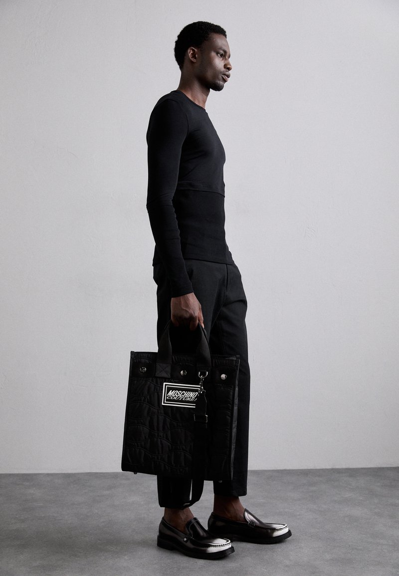 Black quilted tote bag with dual handles and a notable logo patch. Paired with a fitted black long-sleeve shirt and tailored pants.