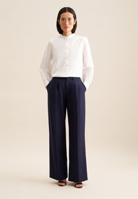 Camicia bianca a maniche lunghe con colletto alto e chiusura a bottoni abbinata a pantaloni navy a gamba larga; tessuto liscio con dettagli a pieghe.