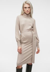 Μπεζ πλεκτό φόρεμα με περιλαίμιο τύπου φουντούκι, μακριά φουσκωτά μανίκια, ζώνη που δένει στη μέση και εφαρμοστή σιλουέτα, φτάνοντας λίγο κάτω από το γόνατο.