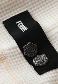 Black fabric label with "FORNER" text above two silver rose-shaped studs with detailed texture, resting on a cream-colored textured surface.
