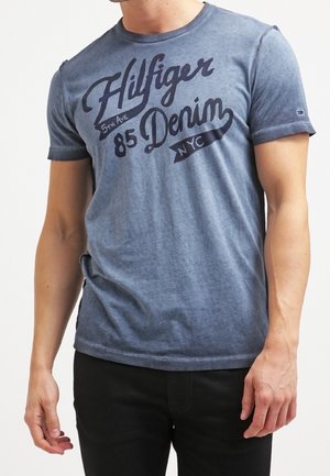 Mężczyzna w wypranej niebieskiej koszulce z krótkim rękawem z napisem „Hilfiger 5th Ave 85 Denim NYC” i czarnych spodniach.