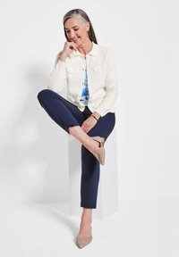 Lichtromige button-up jas over een blauwe bloemetjes top, gecombineerd met donkerblauwe broek en beige puntige schoenen. Glad textuur en slim fit ontwerp.