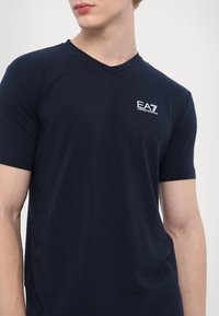 Námořnická modrá polokošile s výstřihem do V vyrobená z jemné bavlny, s malým bílým logem EA7 na levé hrudi a krátkými rukávy.