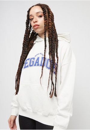 Jeune femme aux longs cheveux tressés, portant un sweat à capuche blanc avec le texte bleu "LEGADO", debout devant un fond uni.