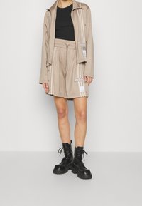 Beige tracksuit jacket with white stripes and logo, paired with matching shorts. Black combat boots complete the outfit.