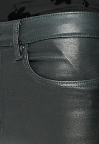 Σκούρο πράσινο, ελαφρώς γυαλιστερό τζιν παντελόνι με μπροστινή τσέπη και κλασικές λεπτομέρειες ραψίματος. Το ύφασμα έχει λεία υφή.