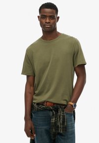 Oliwkowa bawełniana koszulka z okrągłym dekoltem i krótkimi rękawami, noszona z niebieskimi dżinsami i koszulą w kratę zawiązaną wokół talii.