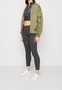 Kvinna som bär en olivgrön bomberjacka över en svart crop top och svarta leggings med rosa tryck, parat med vita Nike-sneakers.