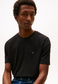 Črna bombažna majica z okroglim izrezom in kratkimi rokavi, z majhno logotipno nalepko v rdeči, beli in modri barvi na prsih.