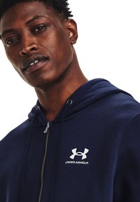 Tumši zilās krāsas zip-up kapuce izgatavota no mīkstas auduma, ar regulējamu kapuci un baltu Under Armour logotipu uz krūtīm.