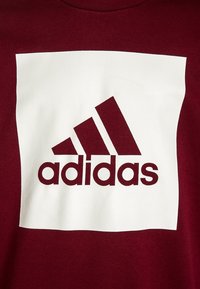 Camiseta color burdeos con un gran gráfico rectangular blanco que presenta el logo de Adidas en burdeos oscuro, exhibiendo un diseño simple y audaz.