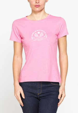 Donna con maglietta rosa a maniche corte con testo e grafica bianchi, abbinata a jeans blu scuro, su sfondo bianco.