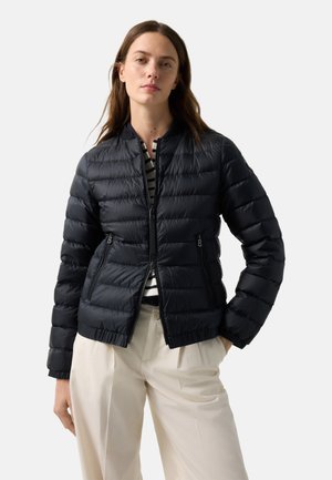 Woman wearing a black quilted jacket over a striped shirt and beige pants, standing with one hand in pocket against a plain background.