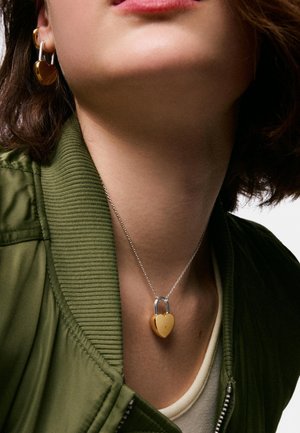 Osoba nosząca złote kolczyki w kształcie serca z zamkiem oraz pasujący naszyjnik z zawieszką na srebrnych łańcuszkach, ubrana w zieloną kurtkę i biały top.