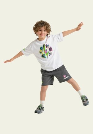 Ragazzo in equilibrio su una gamba con le braccia distese, indossa una maglietta bianca dei Transformers, pantaloncini neri, calze bianche e scarpe da ginnastica grigie.