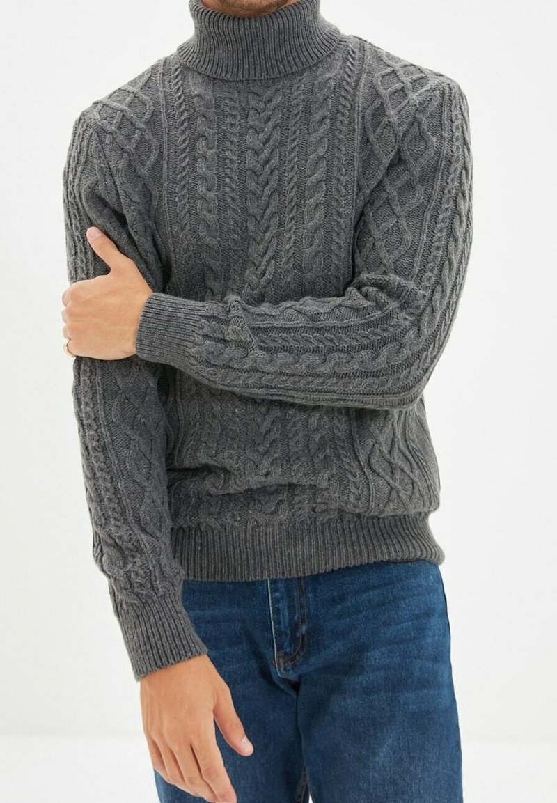 Grauer Kabelstrickpullover mit Rollkragen, gerippten Bündchen und Saumdetails, der strukturierte Muster und eine lockere Passform zeigt. Über blauen Jeans getragen.