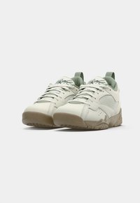 Par de zapatillas bajas blancas y beige con lados de malla y firma en la lengüeta acolchada, colocadas sobre un fondo blanco liso.
