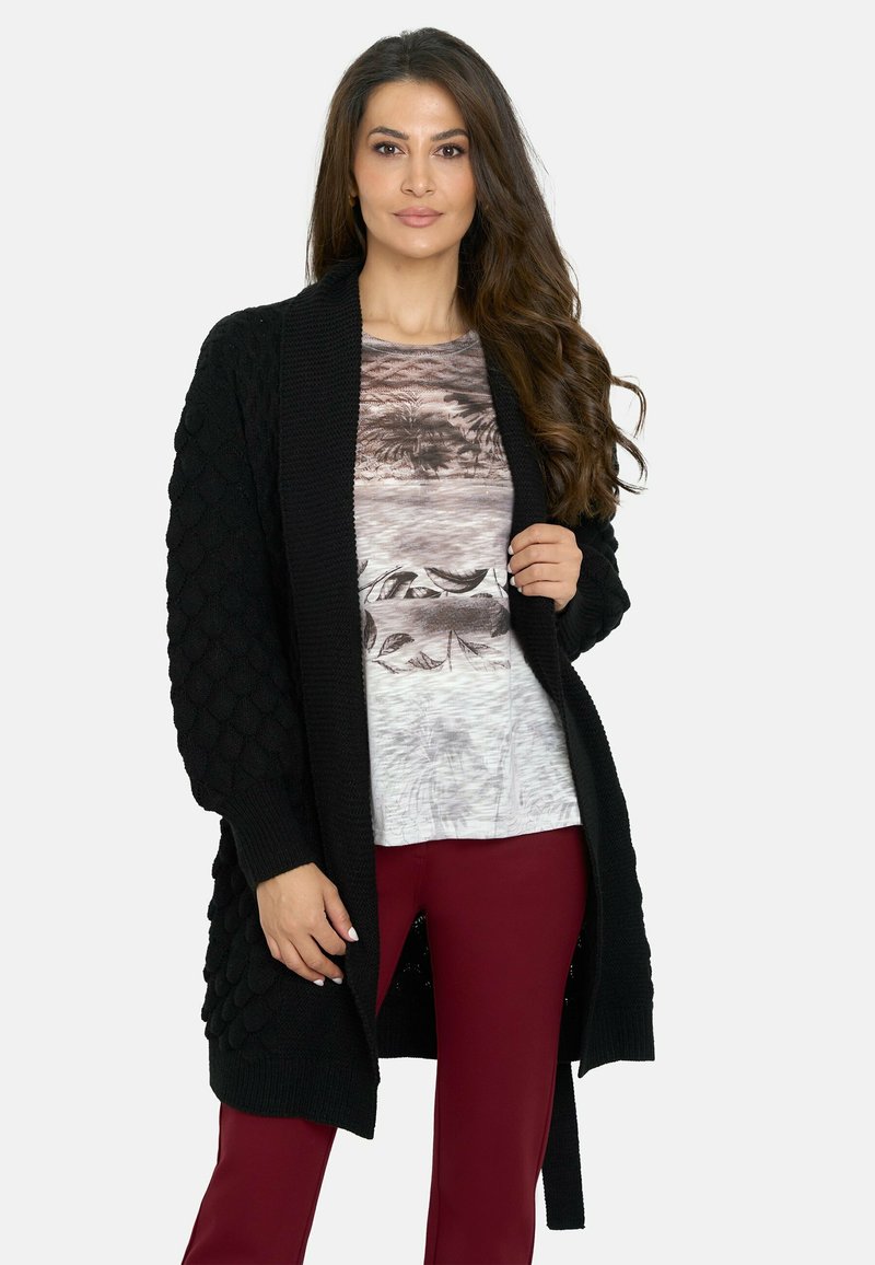 Cárdigan largo negro con patrón texturizado y tejido, con apertura frontal y puños acanalados; usado sobre una blusa clara con diseño escénico y pantalones burdeos.