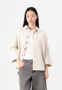 Camicia beige con bottoni e colletto, caratterizzata da dettagli in argento e una vestibilità rilassata. Indossata sopra una t-shirt bianca e abbinata a pantaloni grigi.