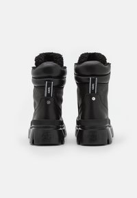 Un par de botas de invierno negras con suelas de goma gruesas, lados de tejido texturizado y cuellos forrados de forro polar, vistas desde la parte trasera sobre un fondo blanco.