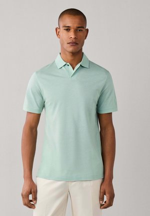 Homme portant un polo manches courtes vert clair et un pantalon beige, debout devant un fond gris uni, face à l’avant.