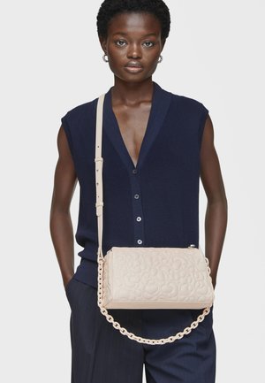 Bolso bandolera beige con patrón texturizado, asa de cadena y correa ajustable. Llevado por un modelo con una blusa sin mangas azul marino contra un fondo blanco.